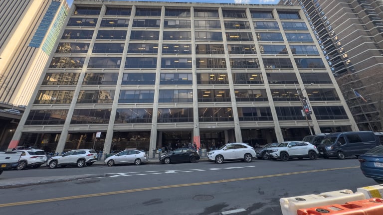Streetview of 100 Gold Street, a large, multi-story residential development with many windows, beside tall skyscrapers. Several cars are parked along the street in front of the building under a blue sky with some clouds.