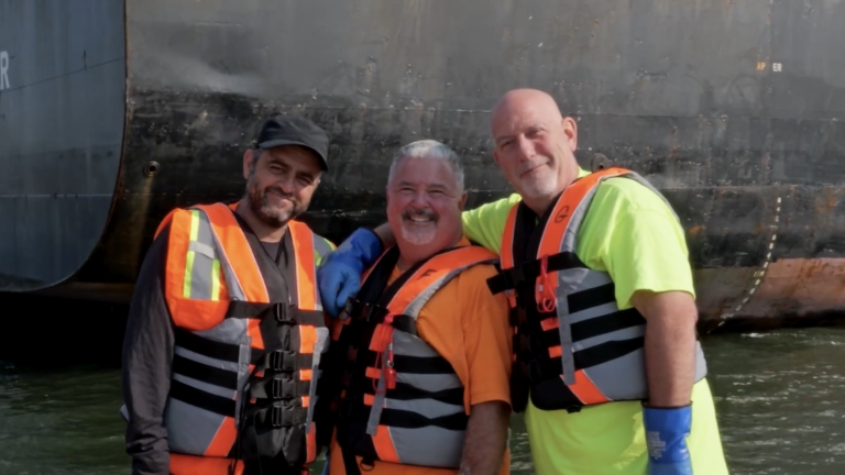 Three smiling men wearing life vests and standing together in front of a large ship on the water.