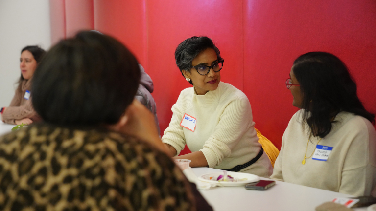Three women sit around a table at a meeting, with two women wearing name tags facing each other while talking.