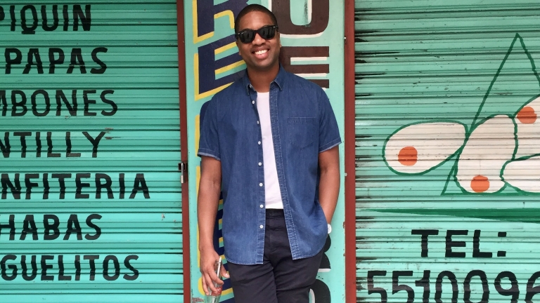 Erich Bilal wearing sunglasses, a blue shirt, and black pants stands smiling in front of a teal shutter with Spanish words painted on it. He holds a drink bottle in one hand. The shutter features abstract shapes and a phone number. Erich Bilal wearing sunglasses, a blue shirt, and black pants stands smiling in front of a teal shutter with Spanish words painted on it. He holds a drink bottle in one hand. The shutter features abstract shapes and a phone number.