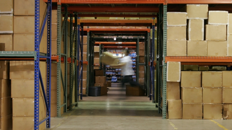 A person in motion, carrying boxes in a warehouse aisle filled with stacked brown cardboard boxes on metal shelves. The floor is concrete, and the shelves extend high, suggesting a large storage facility.