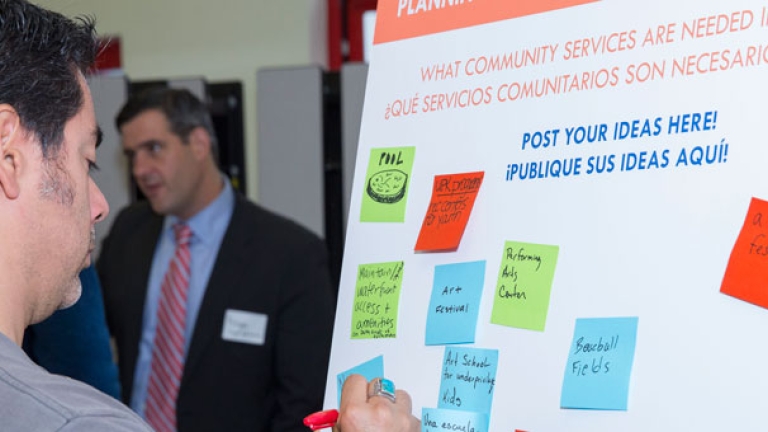 A person writes on a sticky note and places it on a community suggestion board. The board is titled What Community Services Are Needed in Inwood? Several colorful notes, like “Youth Center,” are already posted. Another person looks on in the background.