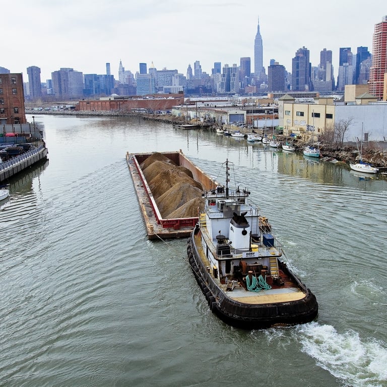 A tugboat pulls a barge carrying sand along Newtown Creek. Surrounding the water are industrial buildings and several small boats. In the background, an NYC skyline with skyscrapers is visible under a cloudy sky. Photo by Mitch Waxman/NYCEDC.