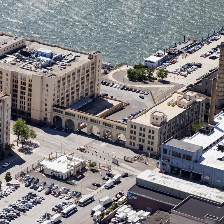 Aerial view of Brooklyn Army Terminal, a waterfront industrial area with large buildings, parking lots, and a long pier extending into the water, with numerous cars parked along the pier. Photo by Albert Vecerka/NYCEDC.