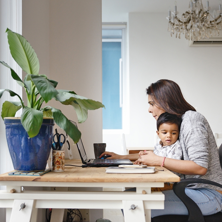 A woman works on a laptop at a wooden desk in a bright room, holding a child on her lap. A large potted plant is on the desk near the window, and a chandelier hangs from the ceiling.