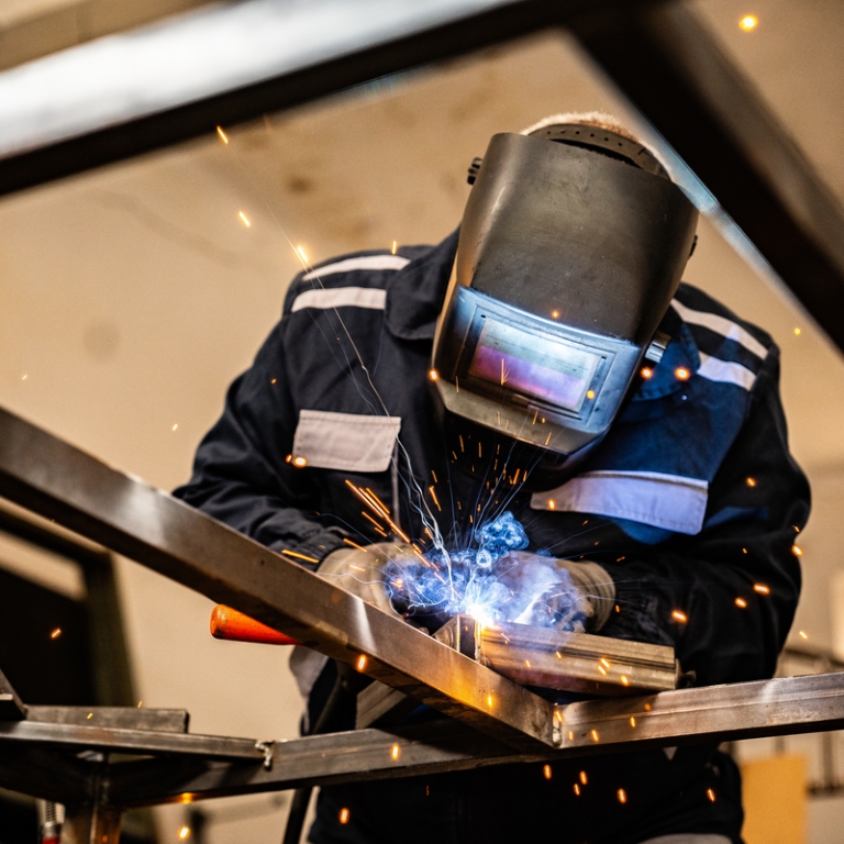 Welder in protective gear working on a metal frame, with bright sparks flying in a workshop setting. Welder in protective gear working on a metal frame, with bright sparks flying in a workshop setting.