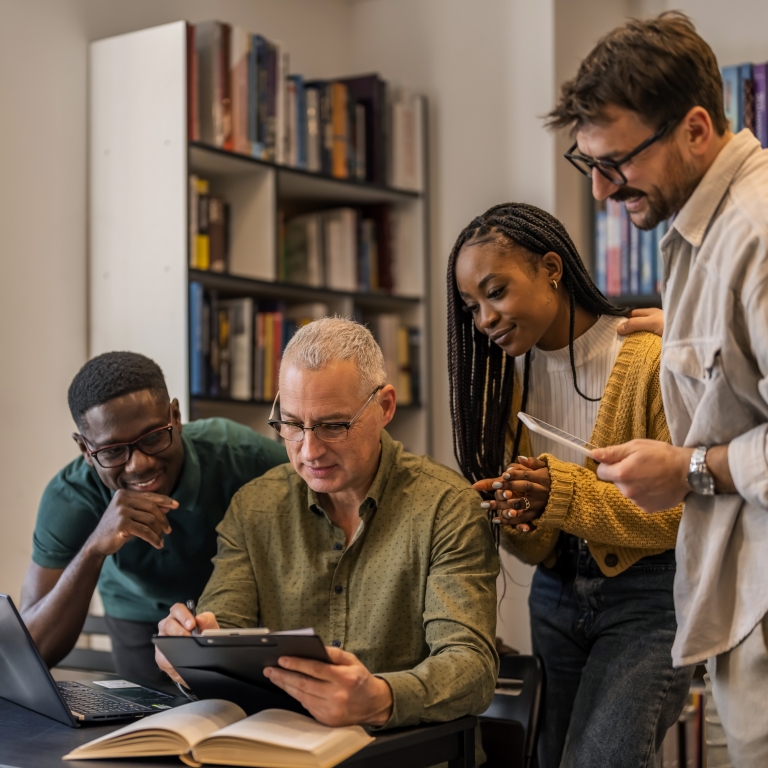 Three students and their professor gather around a table in an office, engaging with a tablet and books, while smiling and discussing.