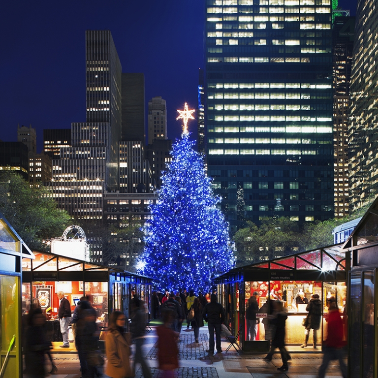 Holiday market in Bryant Park featuring a large, illuminated Christmas tree with blue lights and a star on top. Stalls with various goods line the pathway, while people walk around. Tall, lit-up skyscrapers form the backdrop.