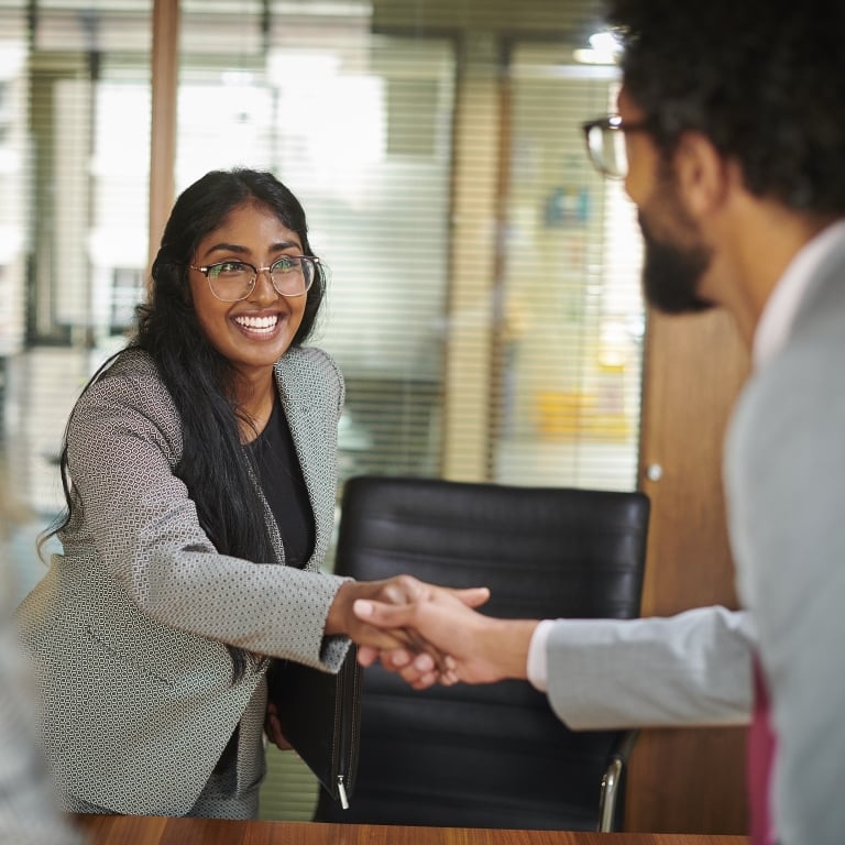 Three people in a meeting room, two shaking hands and smiling, with a focus on the woman in the center.