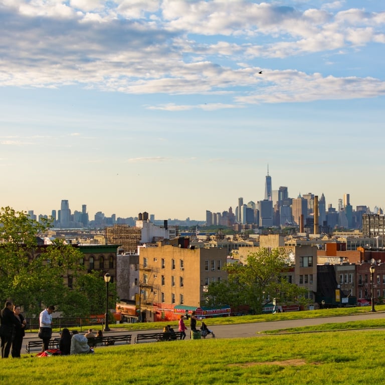 People relax on a grassy hill in Sunset Park with benches, overlooking a New York City skyline in the distance under a partly cloudy sky.