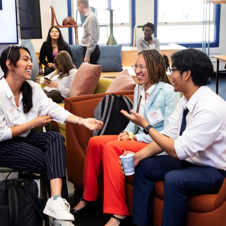 A group of young adults are sitting and talking in a bright, modern office space. They seem relaxed and engaged, with one person holding a coffee cup. In the background, more people are interacting around the room.