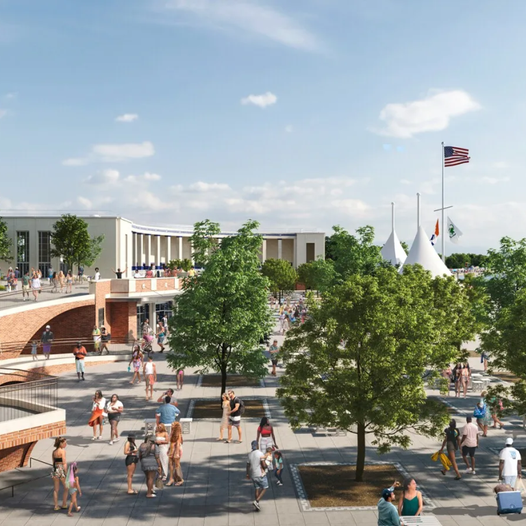 A lively beachside plaza with people walking, sitting, and enjoying the sunny weather. Trees, flags, and modern buildings line the area, while tents and beachgoers are visible near the sandy shoreline and blue water.