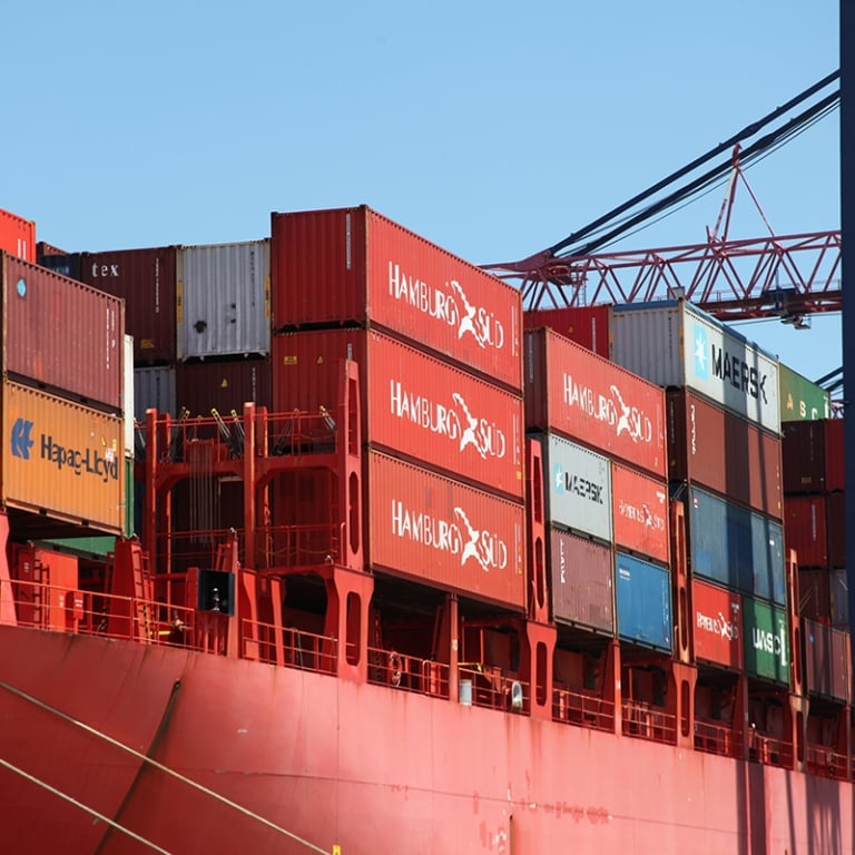 A large red cargo ship loaded with colorful shipping containers is docked at a port, with cranes and blue sky visible in the background.