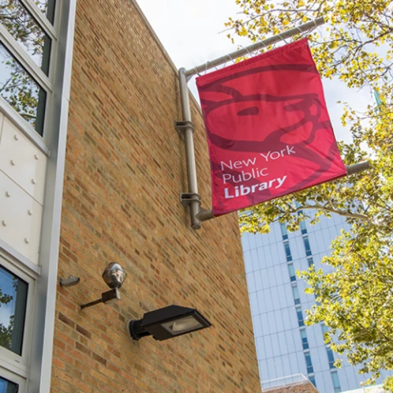 A red New York Public Library banner hangs on a brick building, with trees and a modern glass building visible in the background under a sunny sky.