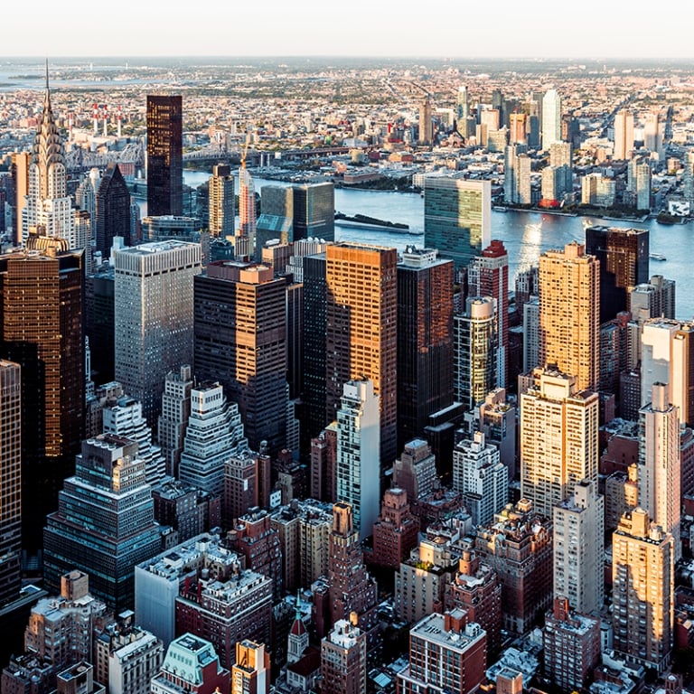 Aerial view of Manhattans dense skyline with numerous skyscrapers, the East River, and part of Queens visible in the background under clear skies.