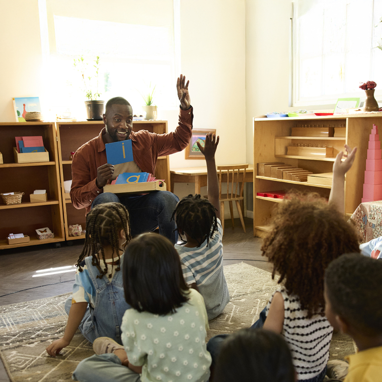 A teacher sits on the floor holding a flashcard and smiles while several young children seated around him raise their hands, eager to answer. The classroom has wooden shelves with learning materials and natural light.
