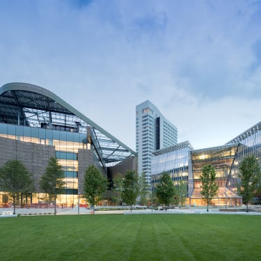 Cornell Tech Campus photo showing modern building complex with glass facades, curved roofs, and a high-rise tower in the background. A landscaped area with trees and a green lawn is in the foreground under a cloudy sky. Photo by Iwan Baan.