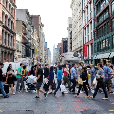 A busy street in SoHo, NYC with numerous people crossing at a crosswalk in different directions. The background features tall buildings, storefronts, and a bus. The sky is clear, and the ground has colorful markings. Photo by Joe Buglewicz/NYC and Company. A busy street in SoHo, NYC with numerous people crossing at a crosswalk in different directions. The background features tall buildings, storefronts, and a bus. The sky is clear, and the ground has colorful markings. Photo by Joe Buglewicz/NYC and Company.