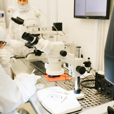 Scientists in cleanroom suits work in a laboratory. One peers into a microscope, while others observe. The lab is equipped with advanced technology, including computers and devices, with a focus on precision and sterility. Scientists in cleanroom suits work in a laboratory. One peers into a microscope, while others observe. The lab is equipped with advanced technology, including computers and devices, with a focus on precision and sterility.