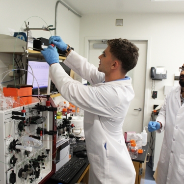Two BiotBat scientists in lab coats, one adjusting equipment attached to a computer while the other observes. The lab is filled with scientific instruments, monitors, and papers. Both are wearing blue gloves, implying an active research environment. Photo by Doug Wongshue. Two BiotBat scientists in lab coats, one adjusting equipment attached to a computer while the other observes. The lab is filled with scientific instruments, monitors, and papers. Both are wearing blue gloves, implying an active research environment. Photo by Doug Wongshue.
