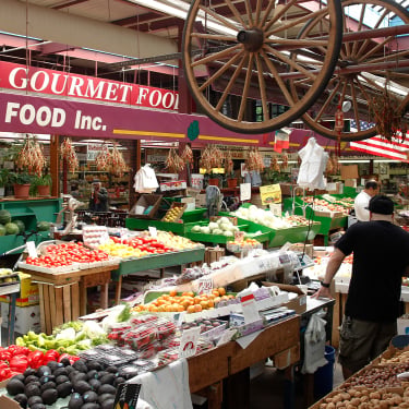 A busy indoor shot of Arthur Avenue Retail Market with various fresh fruits and vegetables displayed on tables. Shoppers browse the produce, and colorful signs hang above the stalls. A large wooden wagon wheel is suspended from the ceiling.