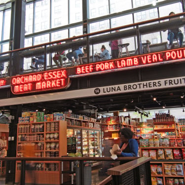 A spacious indoor market with shelves of groceries, neon signs reading “BEEF PORK LAMB VEAL POULTRY” and “ORCHARD ESSEX MEAT MARKET,” and people shopping or sitting on an upper level with large windows.