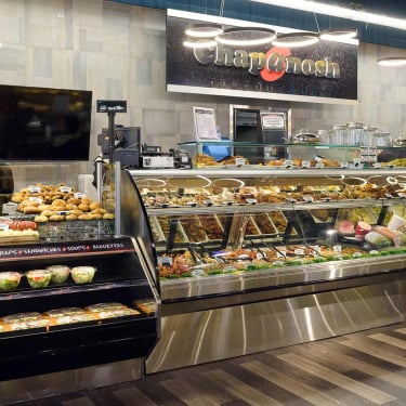 A deli counter displays salads, prepared meals, pastries, and hot foods. Behind the counter, a staff member assists. Signs read Chapnosh and BBQ &amp; Southern Fried Chicken. The floor is wooden, and lighting is bright.