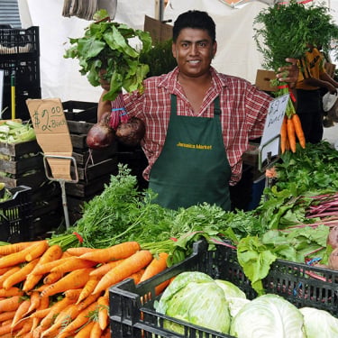 A smiling vendor in a green apron stands behind a market stall displaying carrots, beets, onions, lettuce, and other fresh vegetables, holding bunches of carrots and beets in each hand.