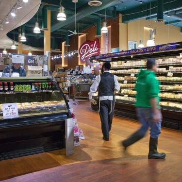 Deli section of grocery store in Certo Market, with prepared foods in glass cases, shelves stocked with salads and dips, and two people walking—one in a green hoodie and boots, another in a vest and apron. Bright overhead lighting.