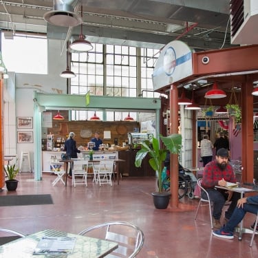 Indoor view of La Marqueta Market with a few people seated at tables. The space has high ceilings, red and orange walls, and greenery. There are various food stalls, and a large window allows natural light to fill the area.