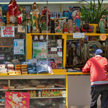 A person in a red jacket stands at a colorful outdoor shop counter filled with religious statues, candles, books, and various spiritual items displayed on shelves and hanging above.