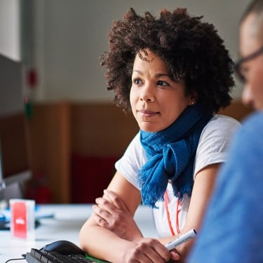 Image promoting Venture Access Alliance. A Black woman with curly hair and a blue scarf is attentively engaging with a man in a blue shirt. They are seated at a desk with computer monitors, suggesting a collaborative work environment.