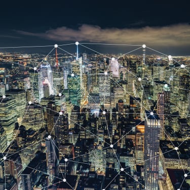 New York City skyline at night with illuminated skyscrapers, overlaid with a digital network grid of interconnected nodes, symbolizing connectivity and smart city technology.