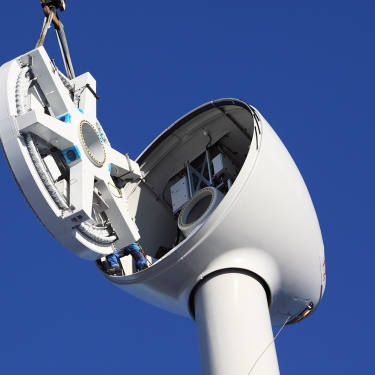 A close-up of a wind turbine being assembled, showing a large crane lifting a mechanical component to the top of the turbine tower against a clear blue sky.