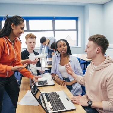 A group of college students in a classroom are collaborating, smiling, and discussing while using laptops. One student stands, holding a 3D-printed wind turbine model, engaging with seated classmates in a bright, modern room.