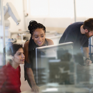 Three people collaborate around a computer screen in a modern office setting with glass partitions. One works at the computer, another watches, and the third stands beside them.