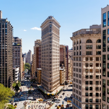 Aerial view of the Flatiron Building in New York City. The triangular building is flanked by other high-rise structures, with streets bustling with vehicles and trees visible in the foreground under a clear blue sky.