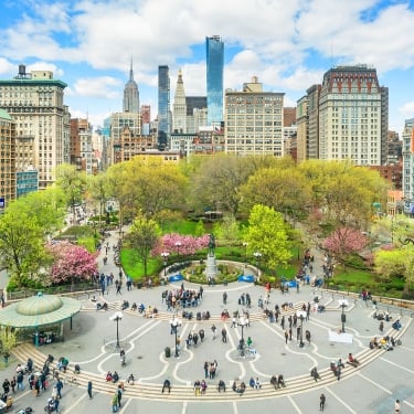 Aerial view of New York's Union Square, a lively urban park surrounded by tall buildings under a blue sky with clouds. The park features lush green trees, blooming pink flowers, a central fountain, and numerous people walking and sitting. Aerial view of New York's Union Square, a lively urban park surrounded by tall buildings under a blue sky with clouds. The park features lush green trees, blooming pink flowers, a central fountain, and numerous people walking and sitting.