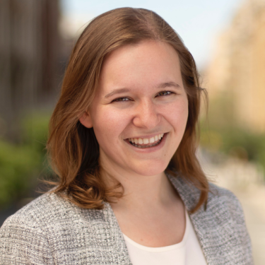 Charlotte Broadbent with shoulder-length light brown hair smiles warmly at the camera. She is wearing a light gray textured blazer and a white top. The blurred outdoor background suggests a city street on a sunny day.