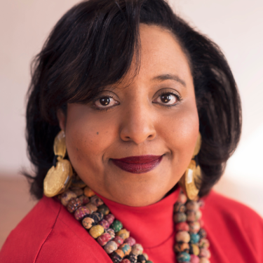 Gayle Jennings-O'Byrne with dark hair wearing a red top, large gold earrings, and a colorful beaded necklace, smiling softly at the camera against a neutral background.