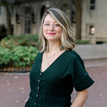 Sydney Mulligan with shoulder-length blonde hair and glasses slightly smiling outdoors. She is wearing a dark green dress with buttons and layered necklaces. The background features greenery and a building with arched windows.