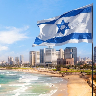 A large Israeli flag waves in the foreground near a beach. In the background, a cityscape with modern buildings lines the shoreline under a partly cloudy sky. The turquoise sea meets the sandy beach with gentle waves. A large Israeli flag waves in the foreground near a beach. In the background, a cityscape with modern buildings lines the shoreline under a partly cloudy sky. The turquoise sea meets the sandy beach with gentle waves.