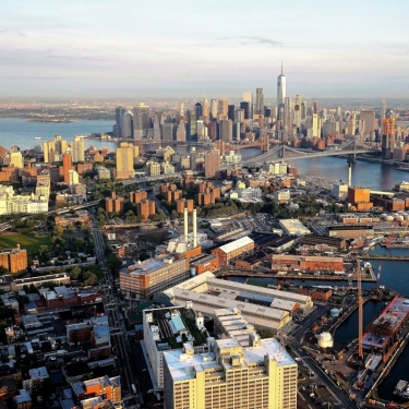 Aerial view of Manhattan, New York City, featuring the dense skyline including One World Trade Center. The Hudson River and various piers are visible, with a mix of urban and waterfront areas under a clear sky.