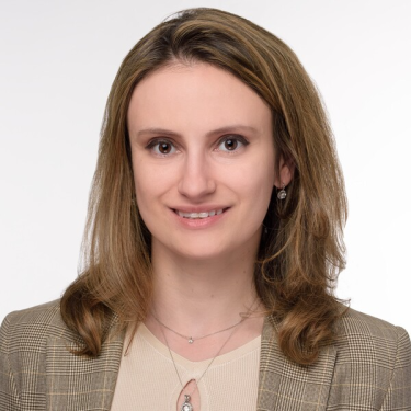 Yana Zorina with light brown, shoulder-length hair is smiling softly at the camera. She is wearing a beige top, a plaid blazer, and a necklace with a small pendant. The background is plain white.