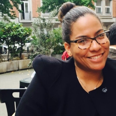 Denis Nolasco with glasses and hair in a bun smiles while sitting outdoors at a café. She is wearing a black jacket and there are trees, plants, and buildings visible in the background.