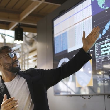 A Black man conducting a meeting with the aid of a large screen. The screen is displaying graphs &amp; data associated with video clips of the earth.