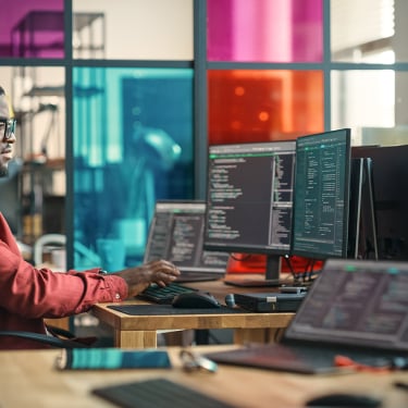 A Black man in a red shirt works at a desk with multiple computer monitors displaying code. The office has colorful glass panels in the background, adding a vibrant atmosphere to the modern workspace.