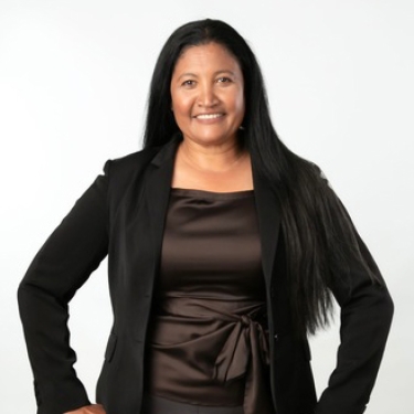 Dr. Corrinne Graham with long black hair wearing a black blazer and a dark brown satin top, standing with one hand on her hip and smiling against a plain white background.