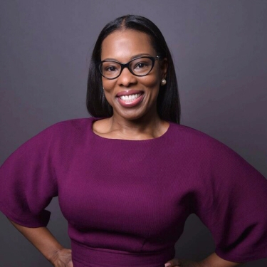 LeShanna Green with long straight hair and glasses smiles confidently at the camera. She is wearing a purple top with wide sleeves and stands against a plain gray background.
