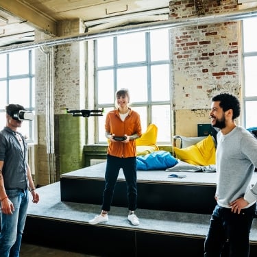 Young start up business people flying a drone inside a big office. One man wearing VR googles and testing new augmented reality software with the drone camera.